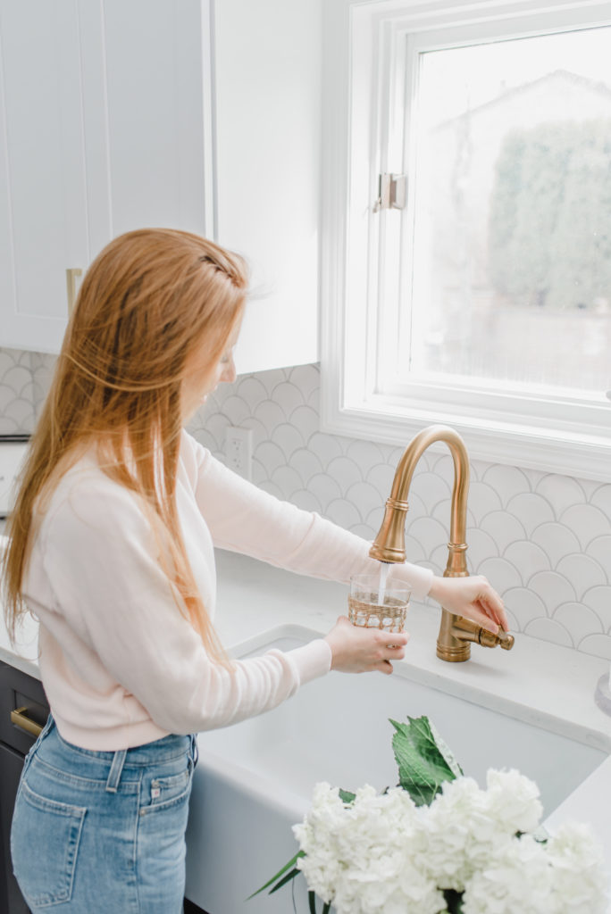 3 Easy Ways To Drink More Water // Filling up a water glass at a farmhouse sink with a gold faucet. There are white hydrangeas in the farmhouse sink and the girl is wearing a light pink sweatshirt and light blue jeans